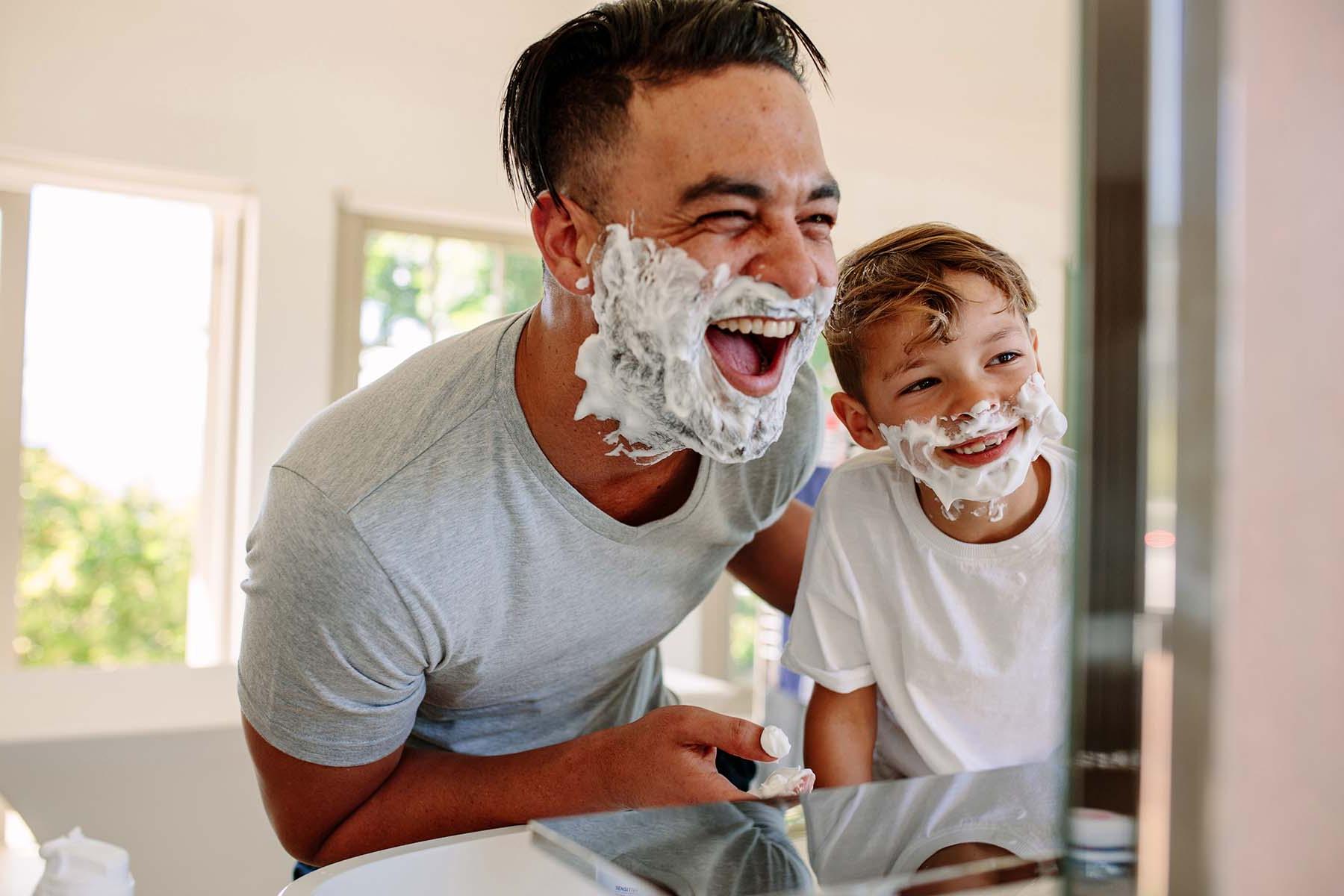 Man and boy with shaving cream on their faces laughing in bathroom.